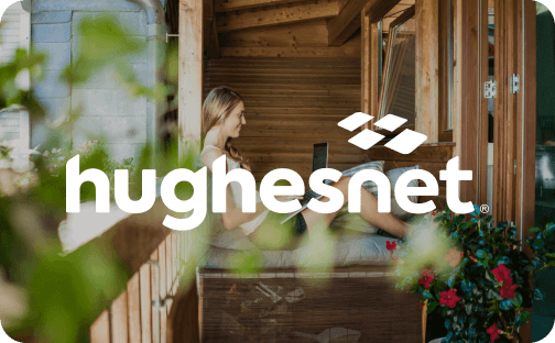 A woman relaxing on a wooden balcony surrounded by greenery, using a laptop. The HughesNet logo is overlaid.